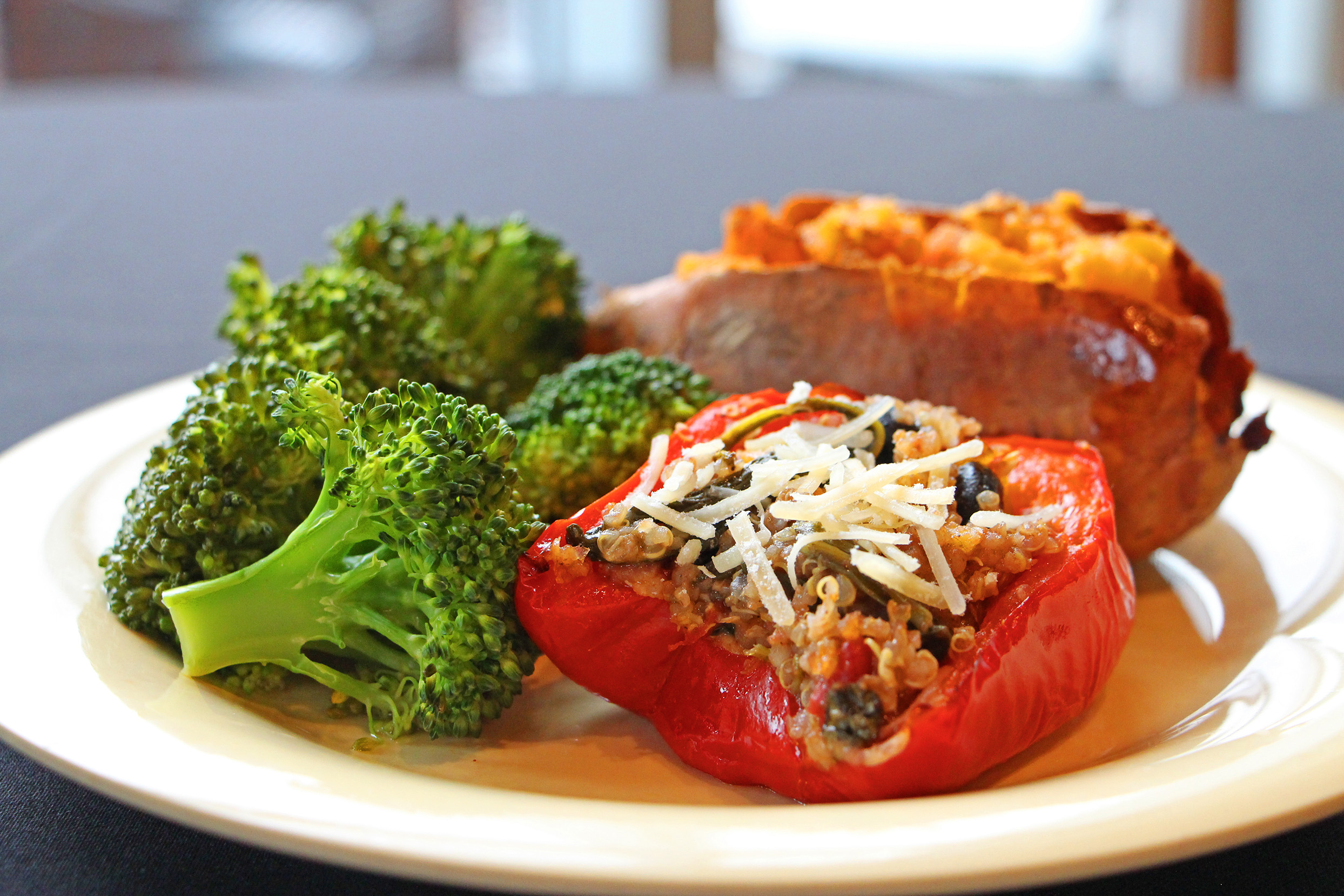 A white plate with steamed broccoli, a sweet potato, and a red bell pepper stuffed with quinoa and cheese.