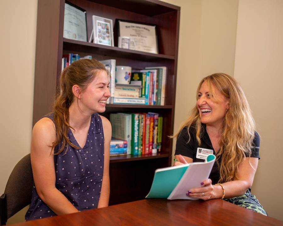 Two people sit at a table talking and smiling. One holds a notebook and points to it. Behind them is a bookcase with nutrition books.